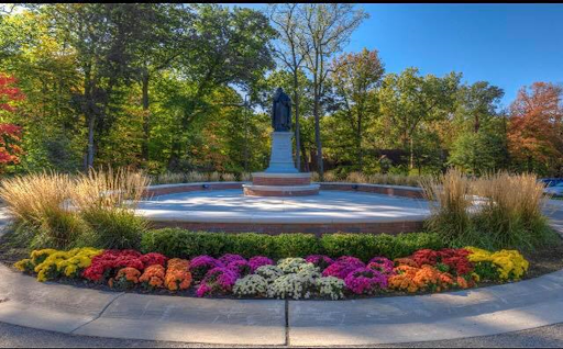 Statue of St. Thomas Aquinas at the center of a paved circle surrounded by a short brick wall lined with bushes, flowers, and grasses. Trees line the backdrop.