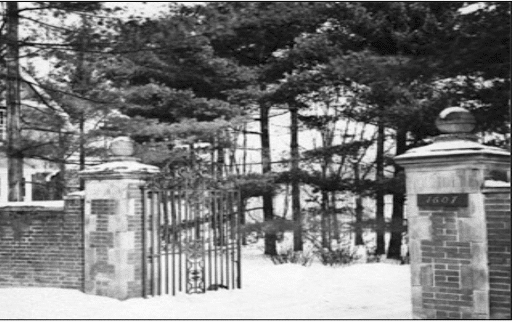 Black and White photo of a wrought iron gate with brick pillars on either side connected to brick walls. Trees fill the space behind the gate, which is open.