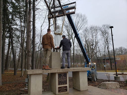 Two people stand on a raised platform guiding the statue of Our Lady of Fatima onto a pedestal. The statue is held by a crane.