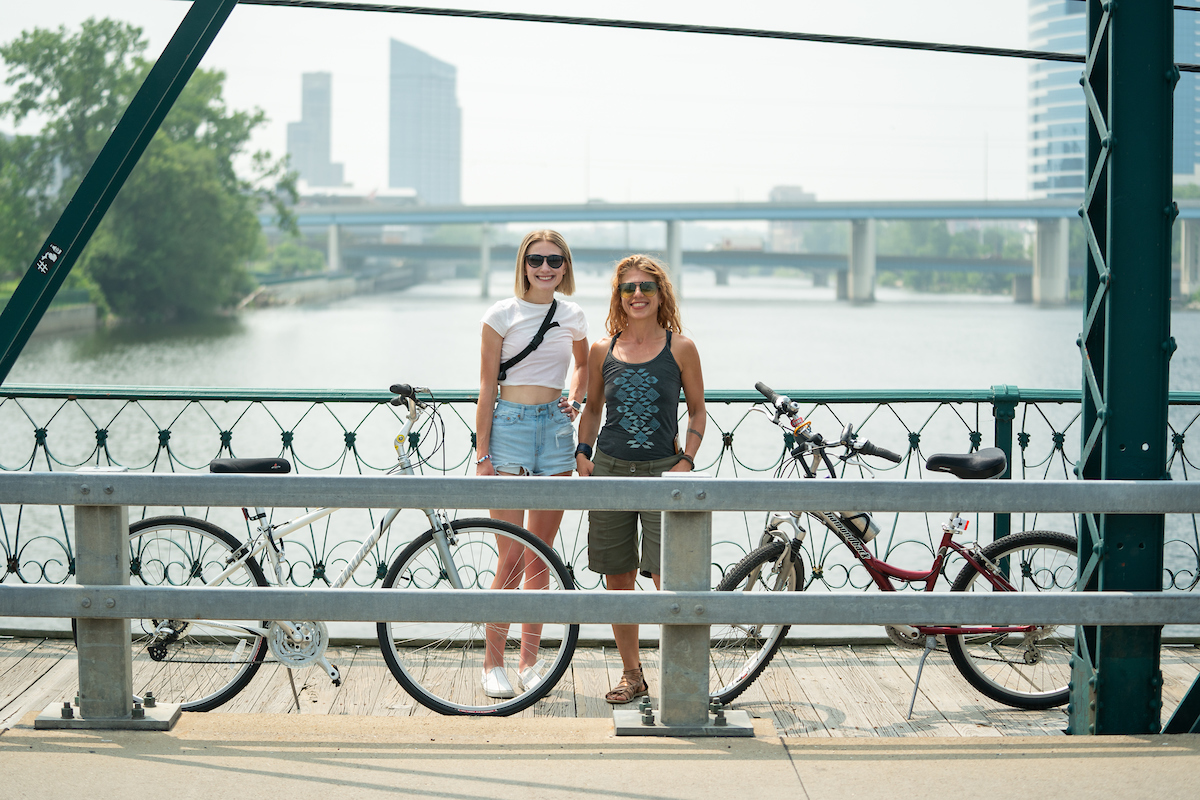 Dr. Lendrum and Arabella Cummings stand on a bridge in Grand Rapids with their bikes, the river and cityscape in the background