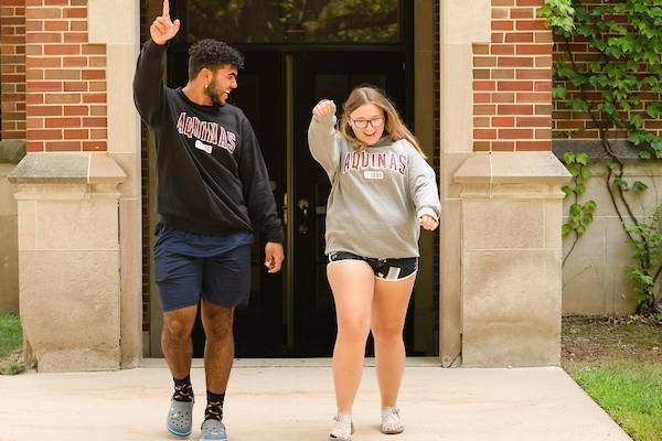 Two students in AQ sweatshirts walk with their hands in the air smiling