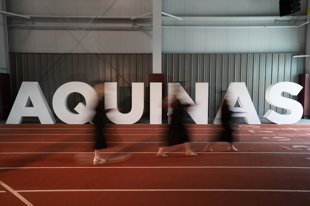 Large Aquinas letters set up on a track with blurred motion students in graduation caps and gowns walking in front