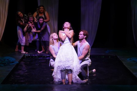 Seven students on stage with water at the center. One student falls back on two with arms dramatically crossed over her chest. A crowd in the back clusters together