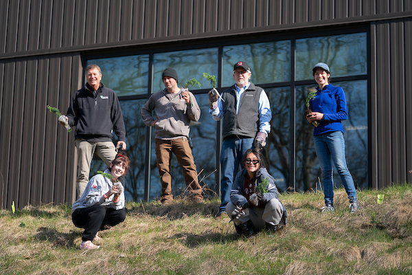 Six AQ community members (staff, faculty, students), smile and hold tree saplings on a hill near Sturrus