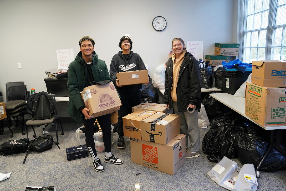 Students smiling holding boxes in a classroom with more bags and boxes stacked in all directions